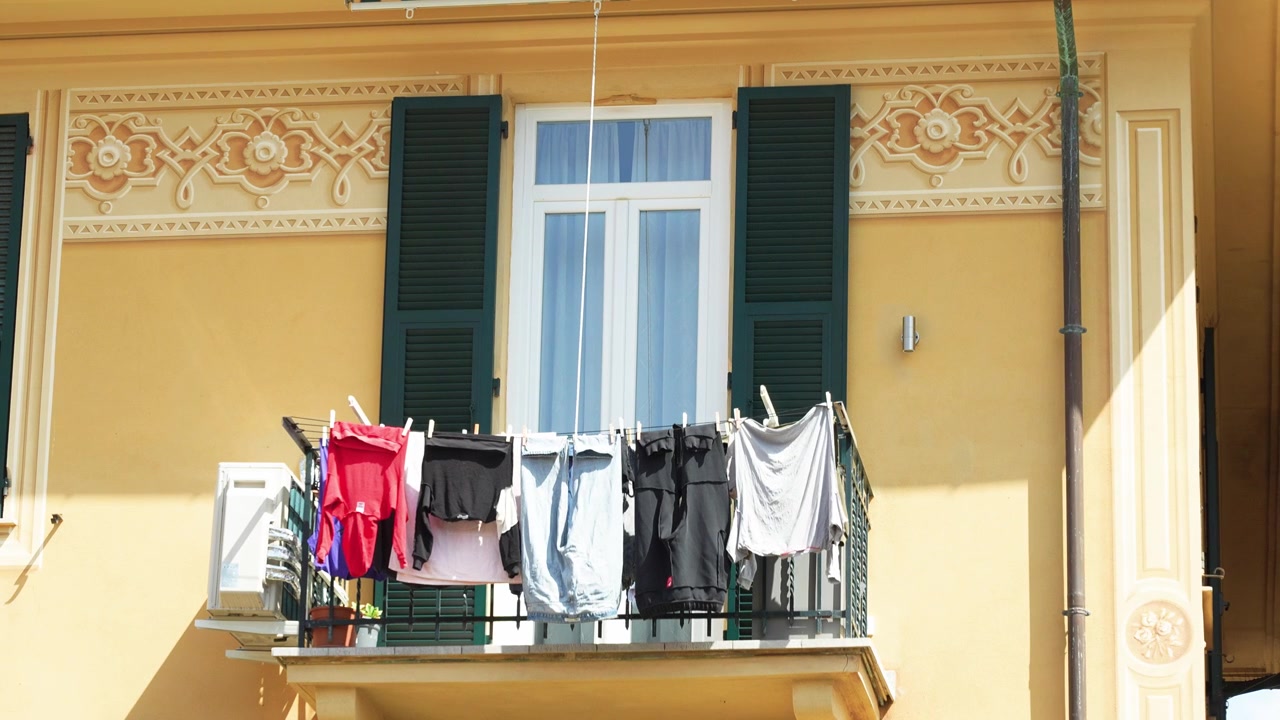 Sunlit balcony adorned with drying laundry on colorful clotheslines, attached to an ornate yellow facade with dark green shutters and decorative accents. Empty, nobody in frame.