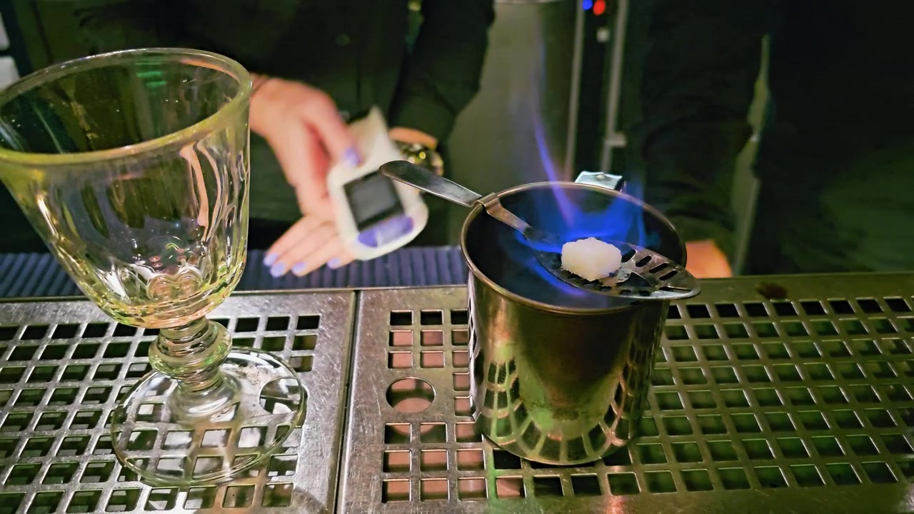 Primo piano di un barista che appoggia un calice vuoto accanto a fiamme blu, un cubetto di zucchero, un cucchiaio forato, una tazza di metallo, un bancone da bar in acciaio inossidabile e l'illuminazione suggestiva di un cocktail lounge, all'interno di notte.