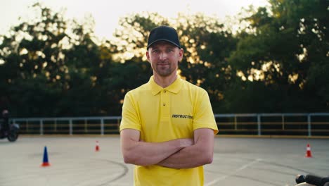 Happy-male-instructor-in-a-yellow-t-shirt-and-cap-folded-his-hands-on-his-chest-and-posing-in-a-motorcycle-school