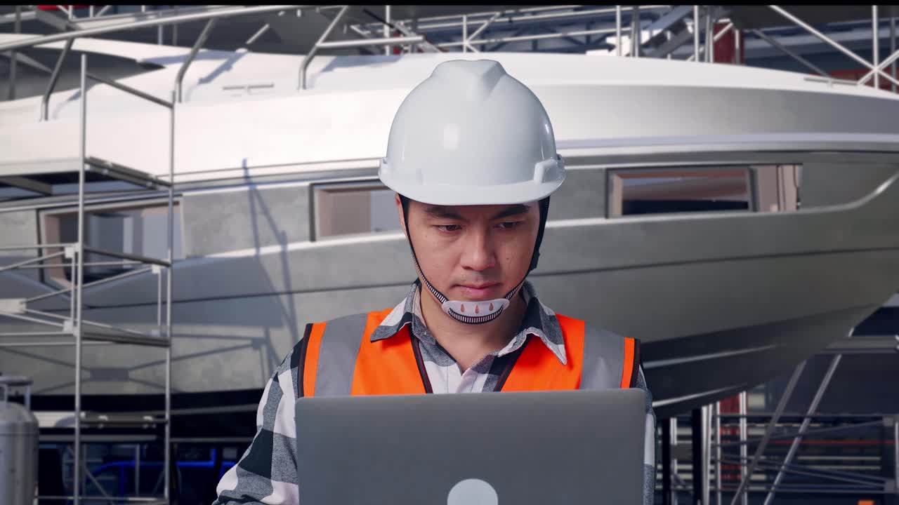 Primer plano de un ingeniero asiático con casco de seguridad trabajando en un portátil mientras está de pie junto a la construcción de un yate en un astillero moderno