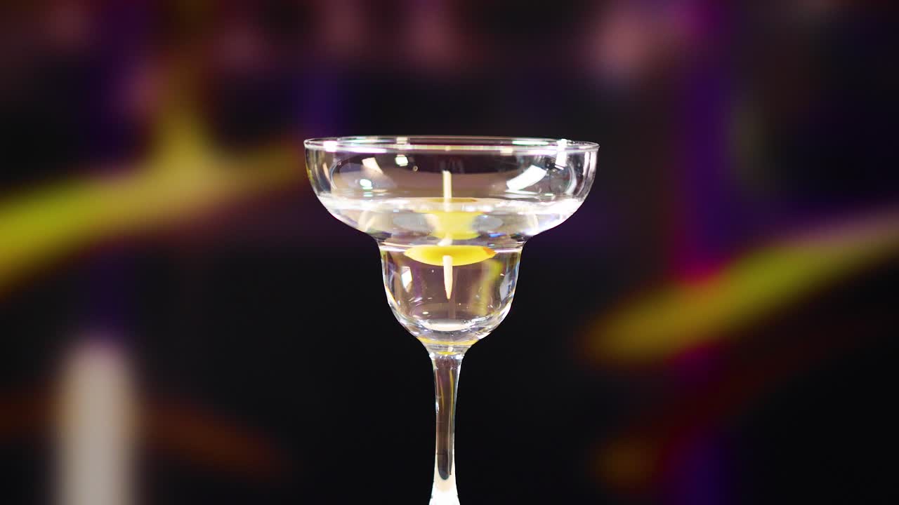 Un twist di limone è posizionato in un bicchiere da martini su uno sfondo vibrante e sfocato. Una luce intensa evidenzia il bicchiere e la guarnizione.