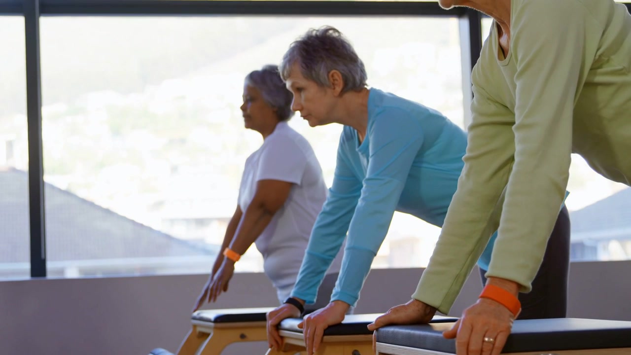 Tres mujeres mayores colocándose en reformadores de Pilates a la señal, inclinándose para el equilibrio de Pilates mientras las alertas se mueven por encima