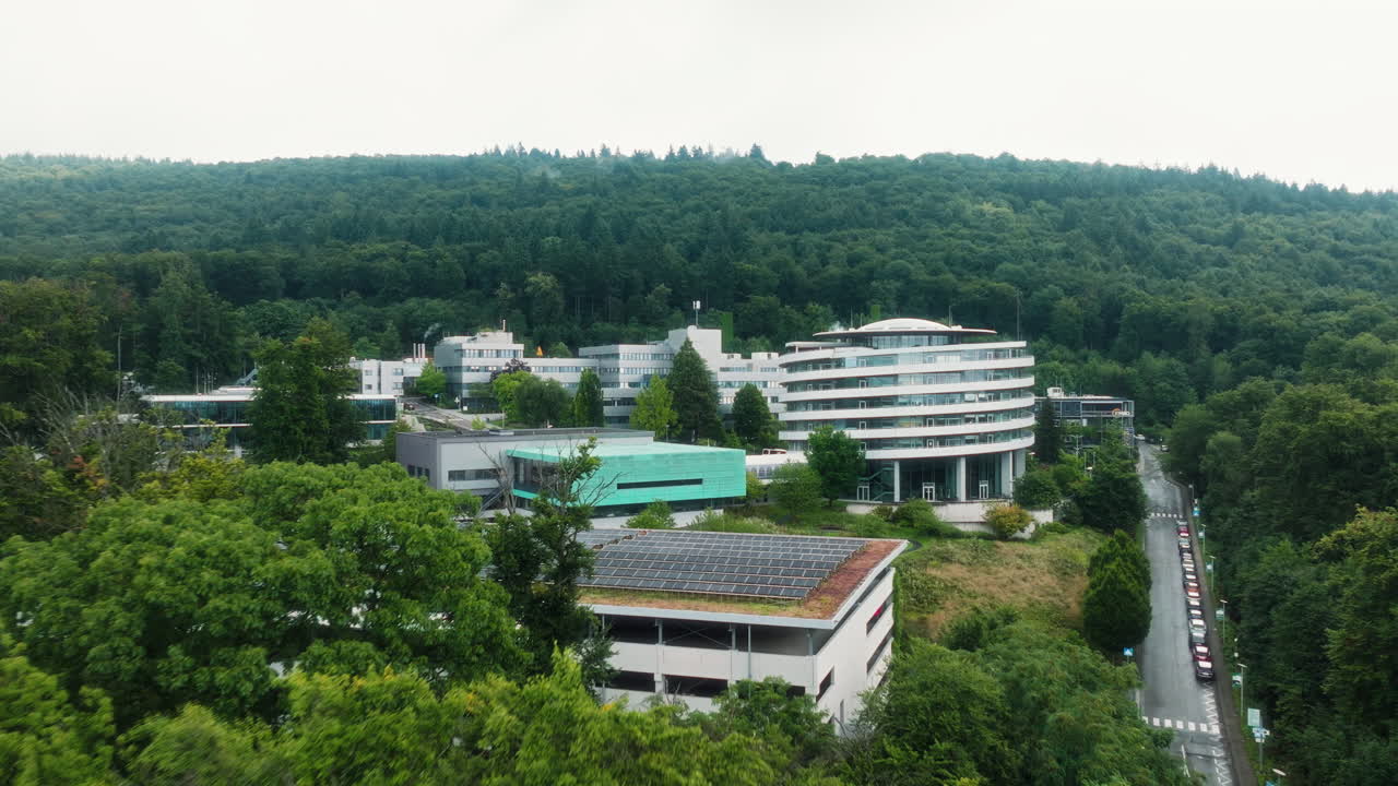 A steady drone shot receding at constant altitude, revealing the DKFZ research campus in Heidelberg, its round building, solar roofs and surrounding forested hills