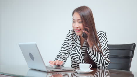 Empleada-Ocupada-Con-Traje-De-Oficina-Usando-Una-Laptop-En-El-Lugar-De-Trabajo-Mientras-Tiene-Una-Conversación-Telefónica-Al-Mismo-Tiempo