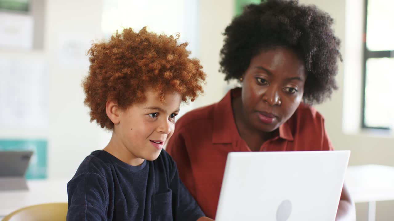 African American woman leaning in, pointing at white laptop guiding school-age boy typing at table