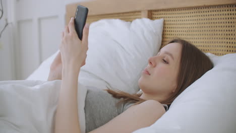 Face-Close-up-on-woman-typing-a-message-on-a-mobile-phone-using-2-hands-while-comfortably-lying-on-a-bed-in-a-hotel-room.-woman-using-smart-phone-and-lying-on-the-bed-in-Morning-after-waking-up