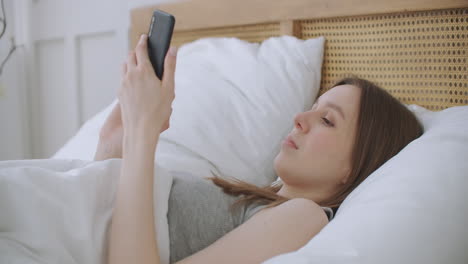 face close-up on woman typing a message on a mobile phone using 2 hands while comfortably lying on a bed in a hotel room. woman using smart phone and lying on the bed in morning after waking up