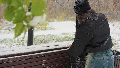 back view of someone opening guitar pack placed on snow-covered park bench, with snow gently falling, a green branch in focus, and a serene winter landscape in the background