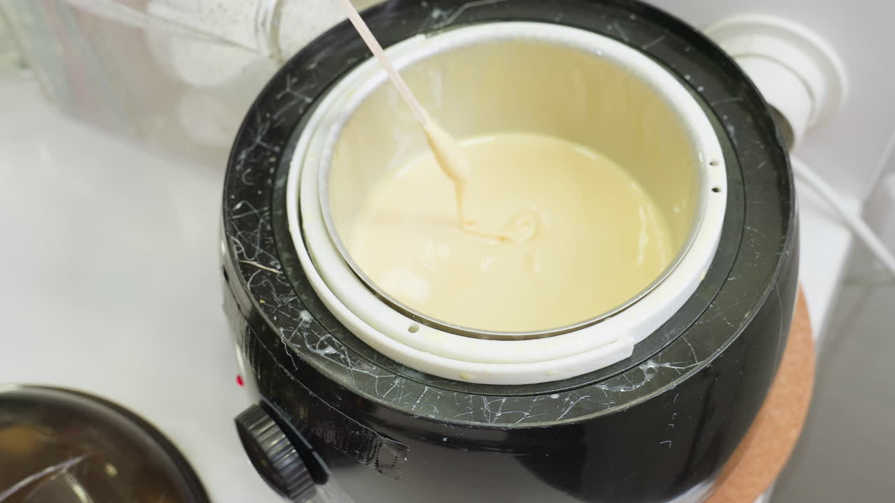 Close up of beautician wearing black glove using wooden stick to stir creamy substance inside electric heater, likely preparing for cosmetic treatment or waxing in professional salon setup