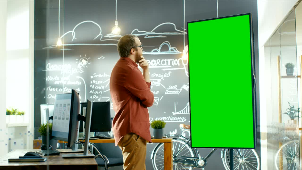 남성 앱 개발자는 모의 녹색 화면으로 인터랙티브 화이트 보드를보고 문제 해결책을 생각하고 있으며, 배경에는 프로젝트 계획이 그려진 칠판 벽을 가진 크리에이티브 사무실이 있습니다.