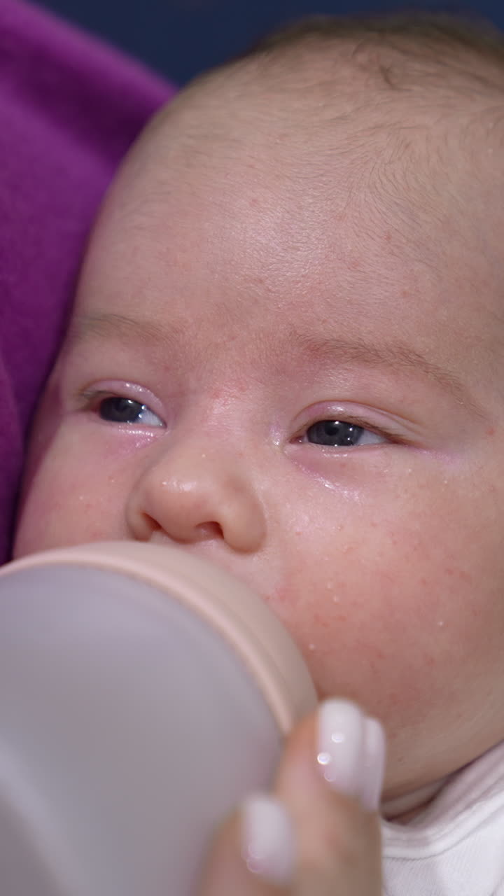Cute infant suckling milk slowly from a bottle. Baby closing eyes and falling asleep while eating. Kid in mommy's hands close up. Vertical video