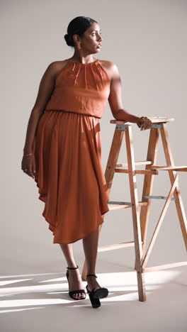 vertical video: elegant woman in stylish dress posing confidently beside wooden ladder in studio