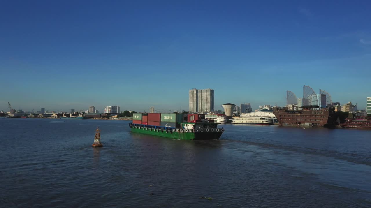 A small Cargo Ship transporting containers of manufactured goods down the Saigon River and Port at Ho Chi Minh City. Landmarks are visible in the background. Drone shot in Sunny  early morning light