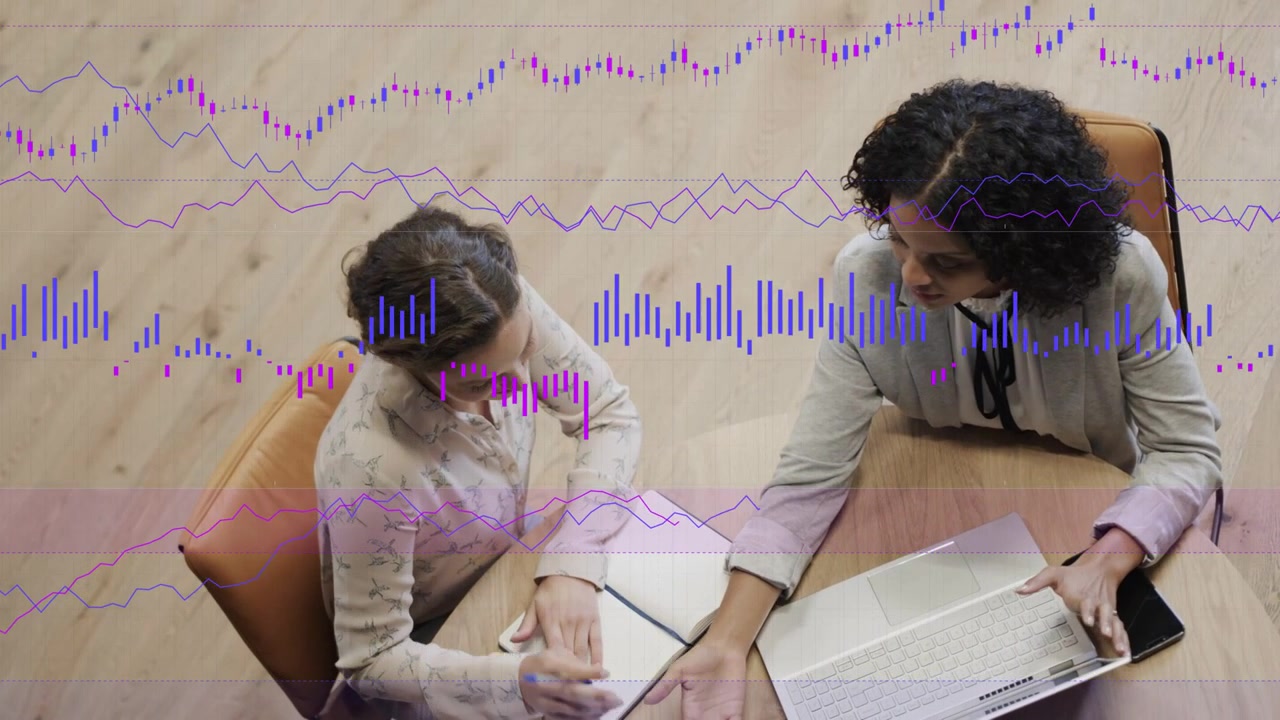 Due donne che si consultano a un tavolo, quella a destra che tocca il laptop e indica, con grafici aziendali che crescono sopra di loro