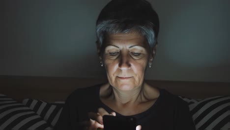 senior woman with short gray hair using a smartphone in a dark room, with the screen light softly illuminating her face, creating a serene and peaceful atmosphere