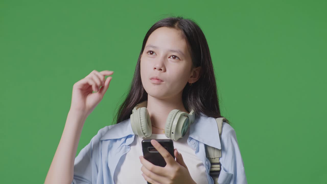 스마트폰을 사용하고 녹색 화면 배경 스튜디오에 서 있는 동안 생각하는 배을 가진 아시아 십대 소녀 학생의 클로즈업
