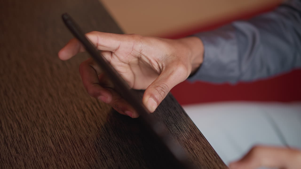 Immigrato che tiene un tablet vicino a un tavolo di legno, con le dita che afferrano il bordo dello schermo in una vista ravvicinata, che indossa una camicia grigia e pantaloni bianchi, ripreso al chiuso durante un uso casuale.