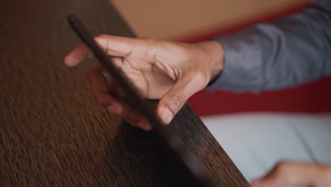 immigrant holding tablet near wooden table, fingers gripping edge of screen in close-up view, wearing gray shirt and white pants, captured indoors during casual use