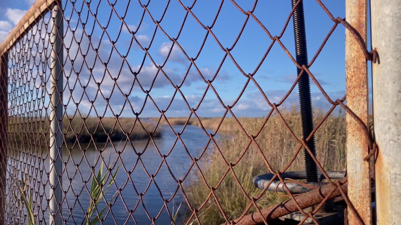 Cerca de arame enferrujada com um mecanismo de cadeado a bloquear a vista de um rio em área húmida e canas secas sob um céu azul, Trebujena, Espanha.