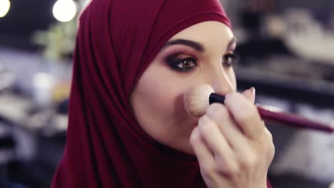 stunningly beautiful girl with traditional arabic purple chiffon hijab covering her head has unrecognizable make up artist's hand applying some blush on her cheeks.