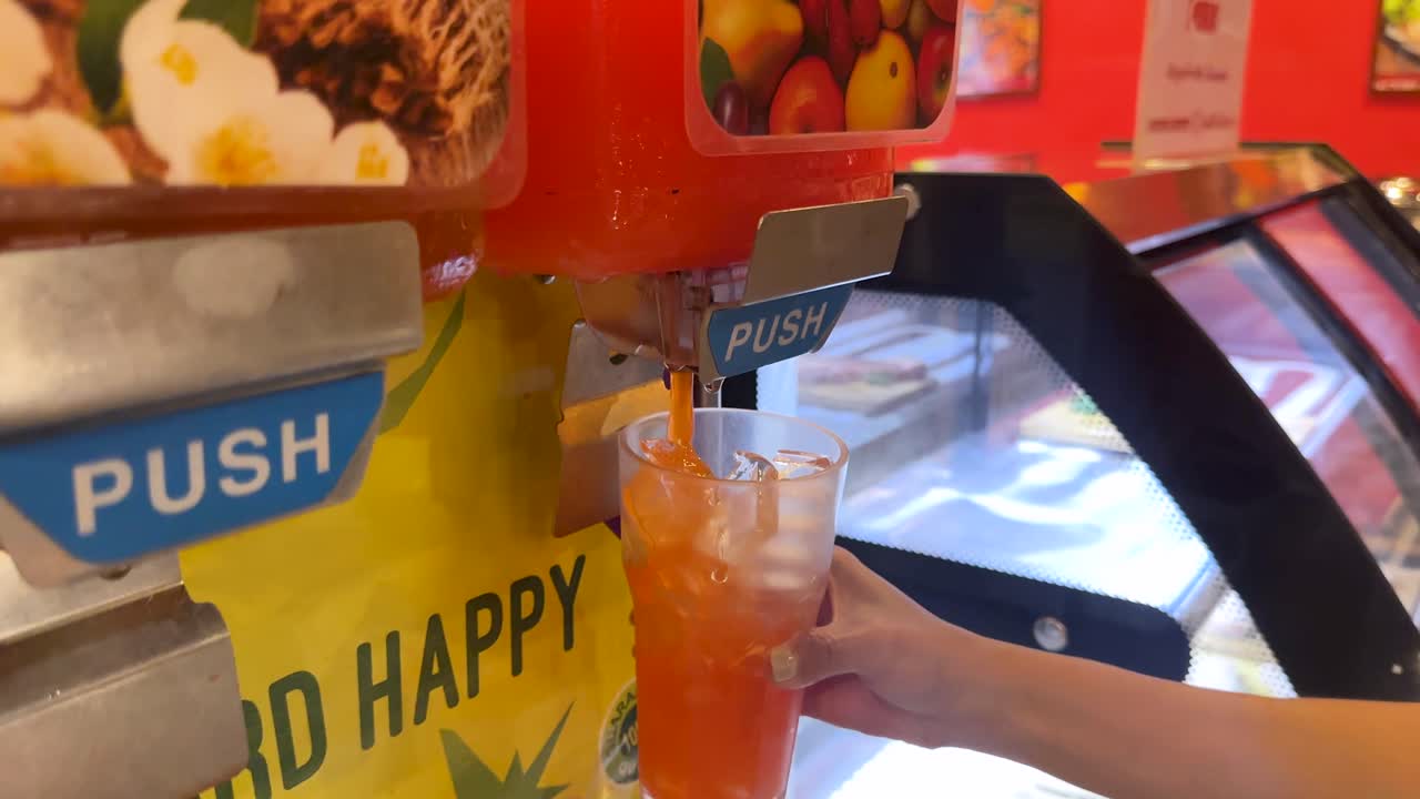 A hand fills a cup with orange drink from a dispenser in a brightly lit Bangkok restaurant