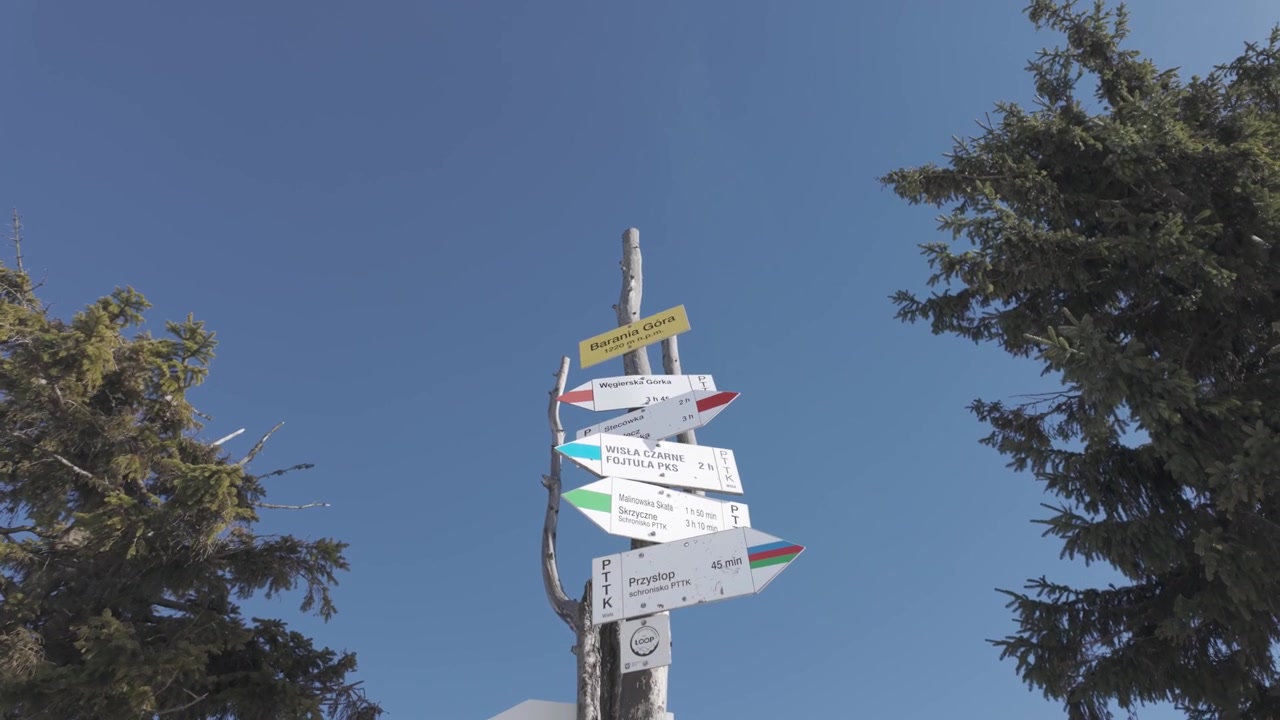 Un cartello segnaletico in legno di montagna con indicatori di sentiero colorati che mostrano percorsi escursionistici e tempi di percorrenza, ripreso lungo un sentiero panoramico all'aperto.