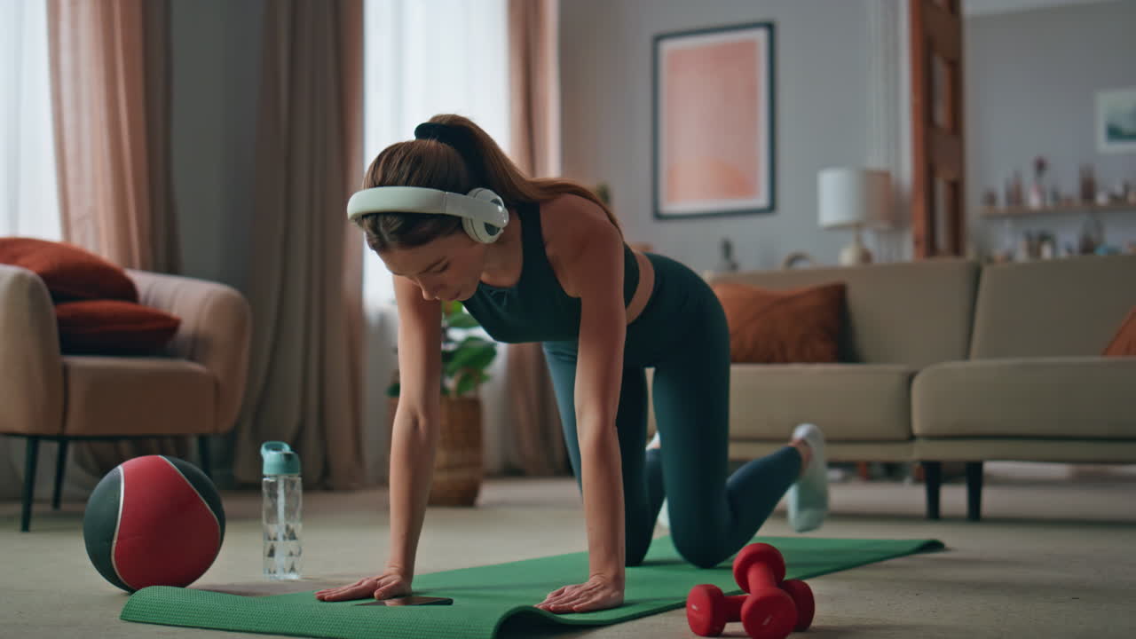 Fitness girl practicing plank listen music in headphones at home. Woman workout
