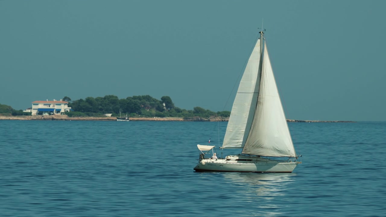 Barca a vela che naviga dolcemente in acque oceaniche calme e blu. Costa distante con alberi verdi e piccole imbarcazioni visibili sotto un cielo sereno, Golfe Juan, Francia.
