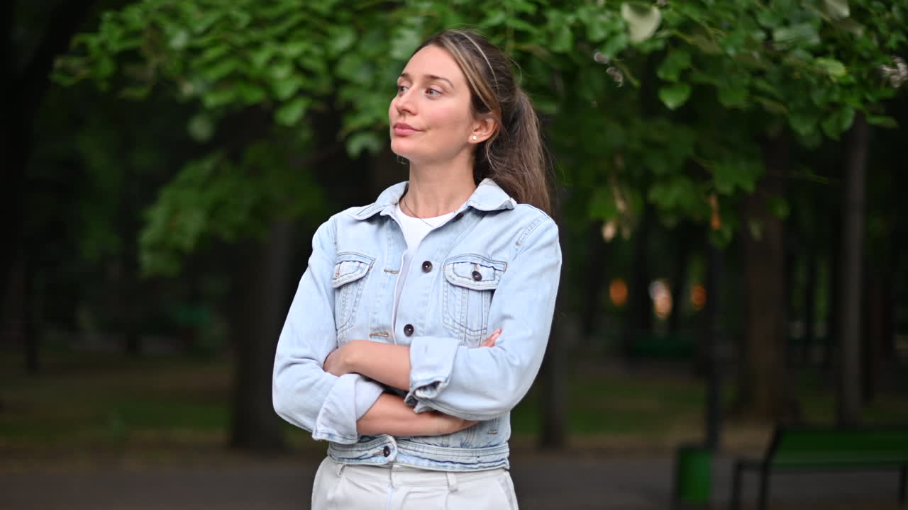 Mujer morena con chaqueta vaquera y pantalones blancos paseando por un parque