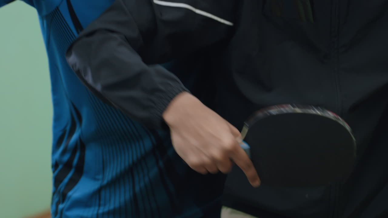 Close up training moment as coach carefully guides player hand holding paddle, demonstrating proper control and movement technique while striking tennis ball during focused indoor practice
