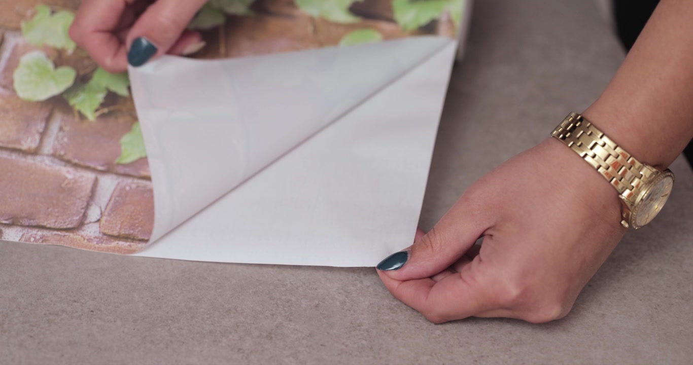 Primo piano delle mani di una giovane donna con un orologio d'oro che stacca una carta da parati adesiva stampata con foglie verdi e motivi a mattoni in 4K.