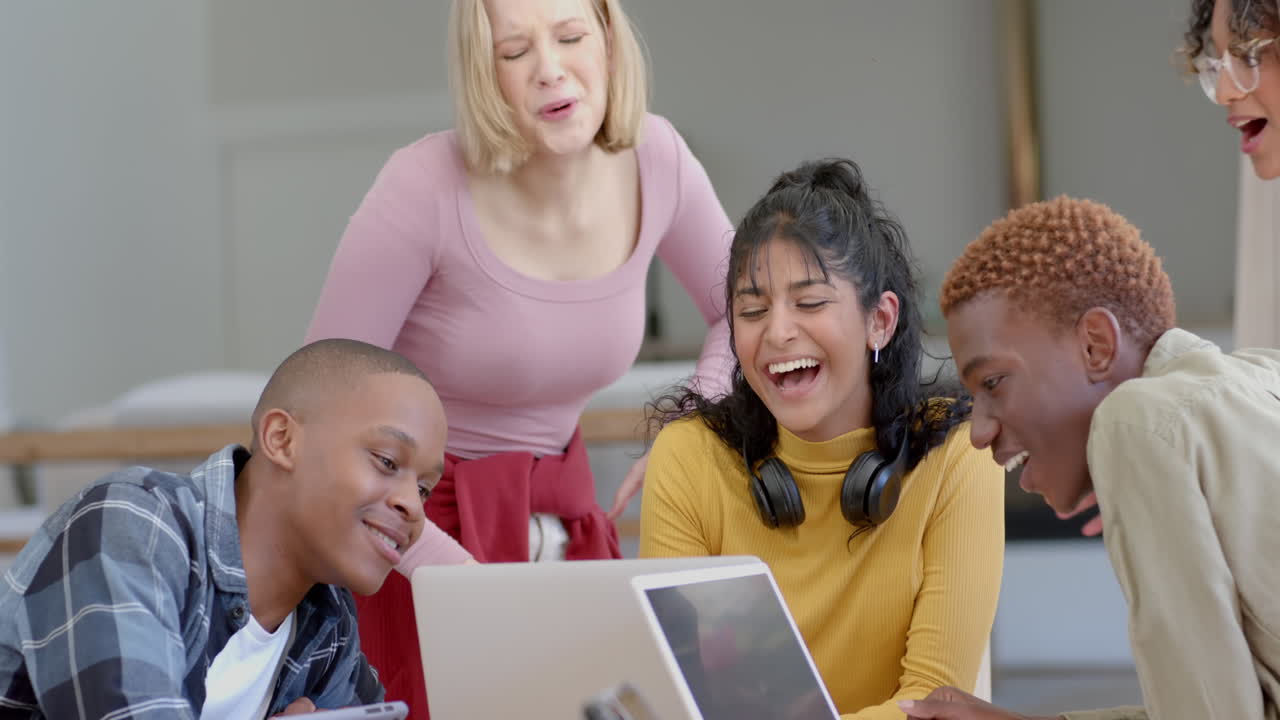 Diverse group of teenage friends studying at table with tablets and laptop at home, slow motion