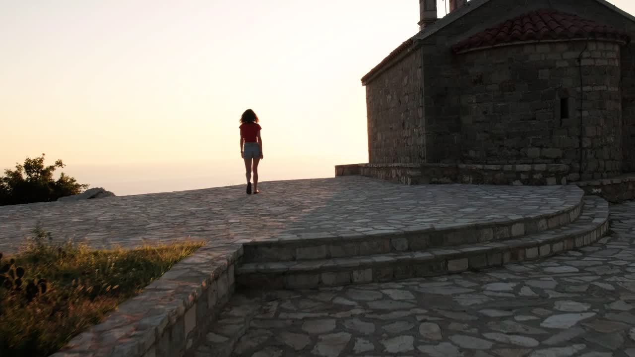 Beautiful lady walking on a viewpoint looking over the Montenegro coastline over top of Sveti Stefan during a beautiful Sunset. Camera walking through a gate following a lady