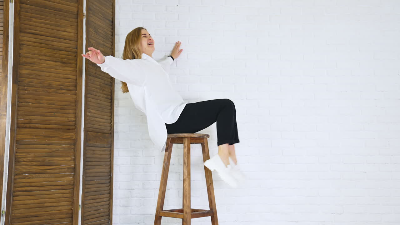 Long-haired lady in white shirt sits on a high stool waving her legs. White modern shoes presentation at the backdrop of white brick wall.