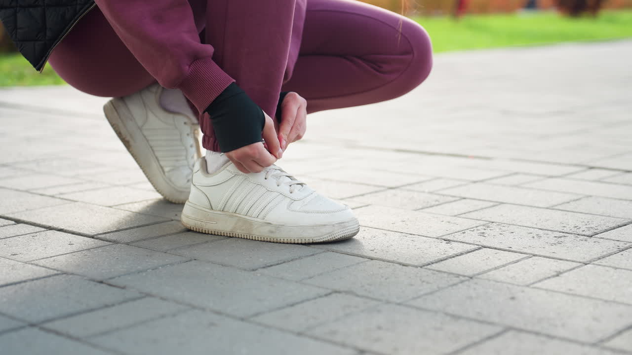 Low angle view urban woman crouching on paved sidewalk tying lace white sneaker before outdoor run near modern apartments wearing maroon tracksuit and fingerless glove while seated on workout bench
