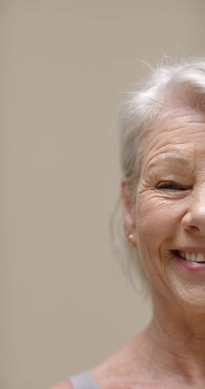 Vertical video: Senior woman smiling, tilting as camera rolling on beige wall, earring, copy space