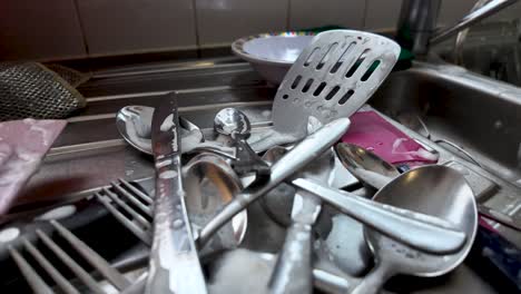 metal cutlery soaking in soapy water in a kitchen sink, ready to be washed. low angle, push forward
