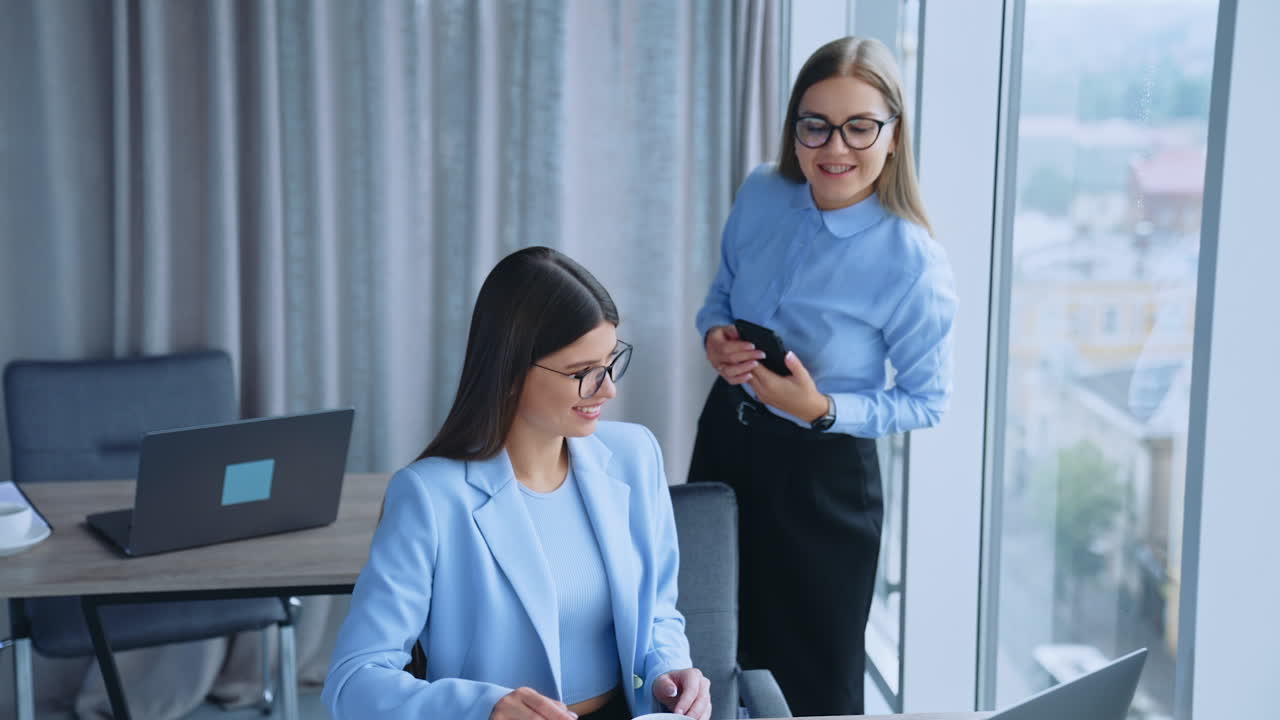 Communication between colleagues at work. Two female employees talking cheerfully and smiling being in the light modern office.