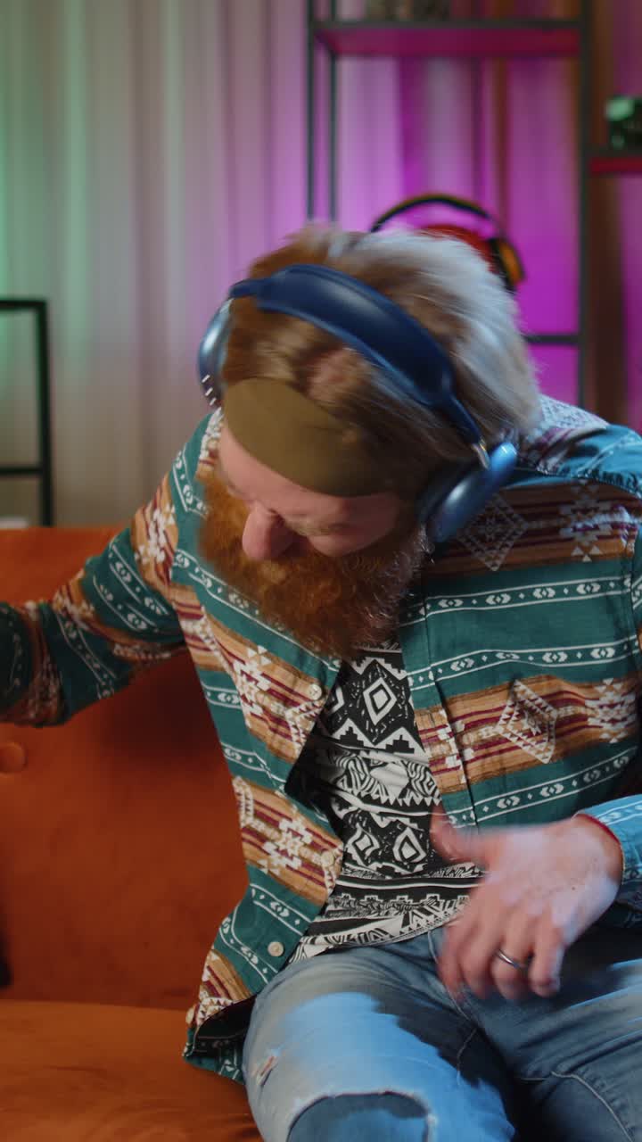 Uomo con le cuffie che balla sul divano a casa ascoltando la sua musica preferita suonando una chitarra immaginaria