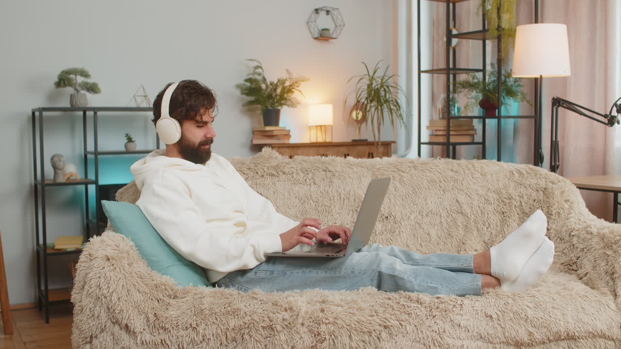 Happy relaxed indian young man in wireless headphones listening to music using laptop on sofa couch