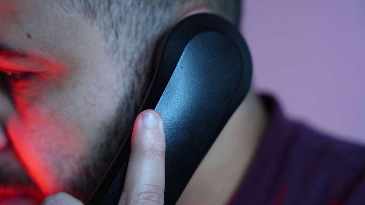 Primer plano extremo de un hombre serio sosteniendo un auricular de teléfono fijo negro en su oído. La dramática iluminación de estudio roja y púrpura resalta su rostro y mano en esta toma cinematográfica de interior.