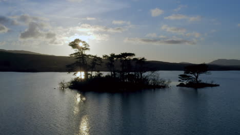 Backlit Lough Bofin’s islands at dusk, golden light reflecting on the calm water and distant hills, static aerial, Connemara, Galway, Ireland