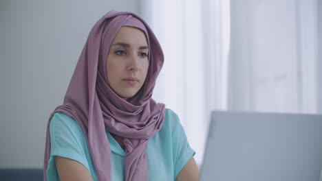 Beautiful-young-muslim-woman-is-working-on-laptop-on-her-workplace.-A-young-muslim-woman-sitting-in-front-of-a-laptop-screen-looks-up-and-looks-at-the-web-camera