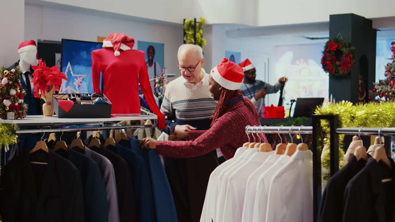 un cliente de edad avanzada recibe ayuda de un asistente minorista en una tienda de ropa decorada de forma festiva para determinar si un elegante blazer es el adecuado. empleado ayudando a un anciano en una tienda de moda adornada de navidad