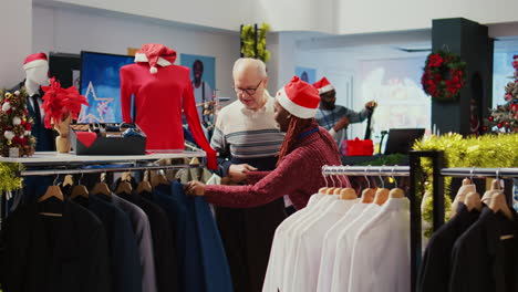 Un-Cliente-De-Edad-Avanzada-Recibe-Ayuda-De-Un-Asistente-Minorista-En-Una-Tienda-De-Ropa-Decorada-De-Forma-Festiva-Para-Determinar-Si-Un-Elegante-Blazer-Es-El-Adecuado.-Empleado-Ayudando-A-Un-Anciano-En-Una-Tienda-De-Moda-Adornada-De-Navidad