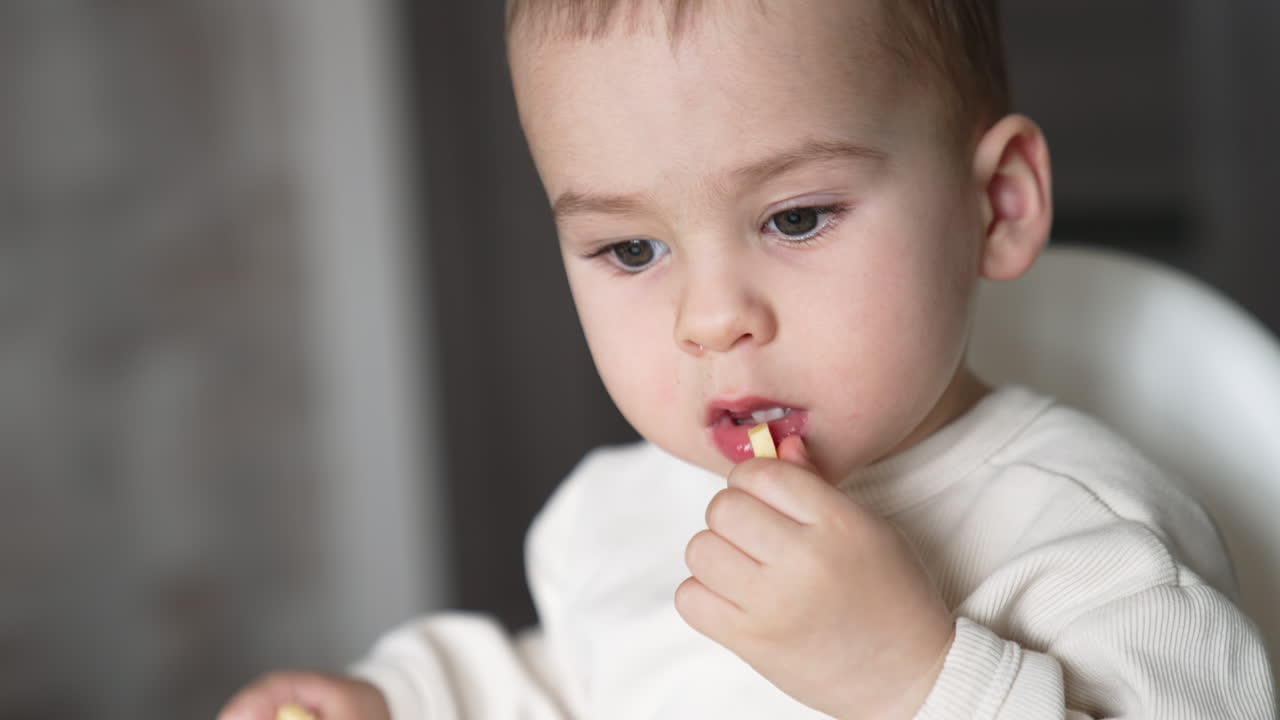 Lovely baby in white sweatshirt takes a piece of cheese from the table. Kid taking food to his mouth thoughtfully. Close up.