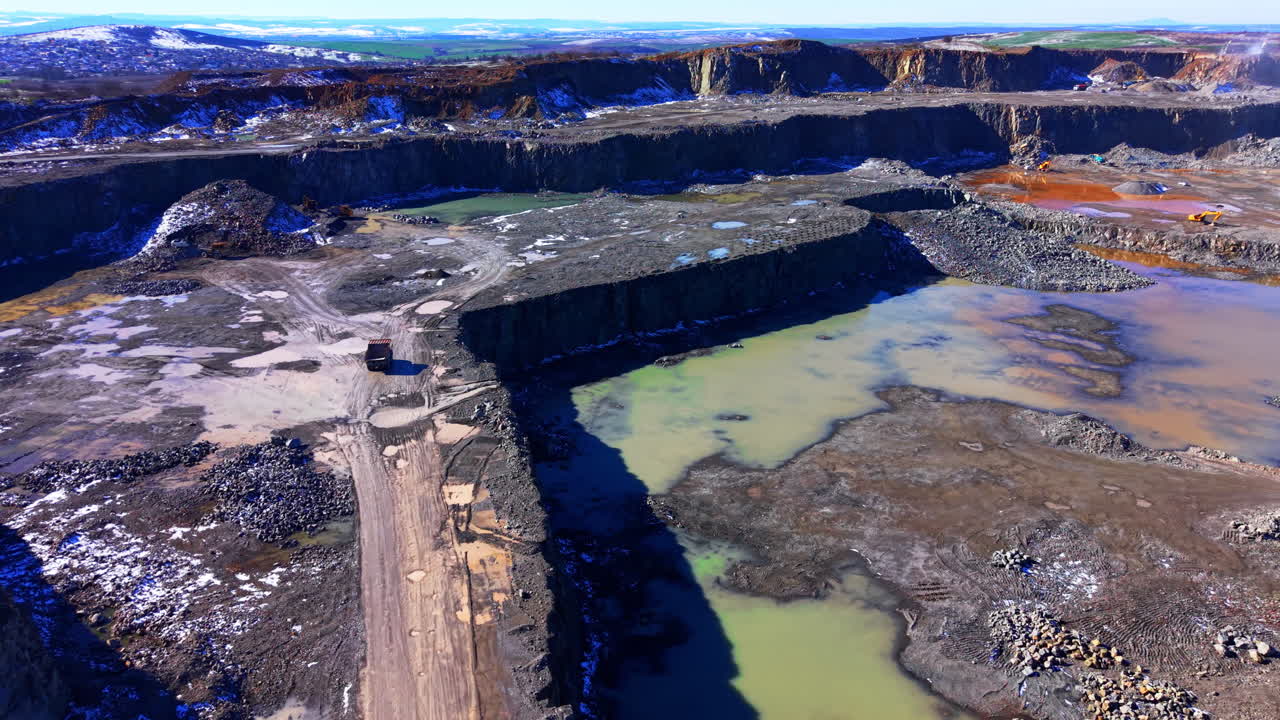 Large scale industrial stone quarry featuring tiered excavation walls, heavy machinery, and pools of mineral-rich water. Wide angle aerial view of open pit stone quarry with water