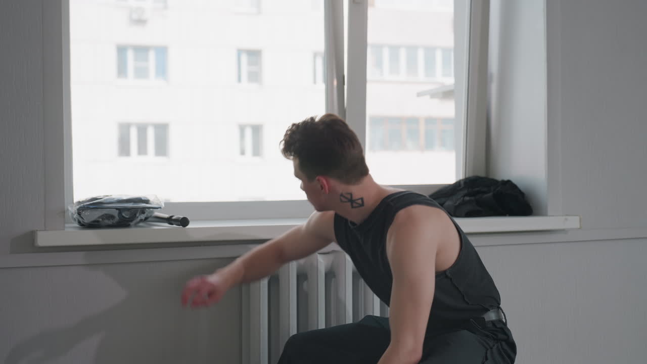 Seated teen in black sleeveless top holds jacket near bright apartment window, examines leather garment before carefully folding and placing it into navy rolling suitcase on floor