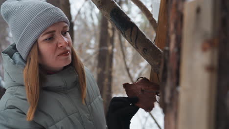 woman in gray beanie and winter coat places piece of bread into wooden bird feeder mounted on snowy tree in forest, focused and caring expression on face, showing compassion and connection with nature