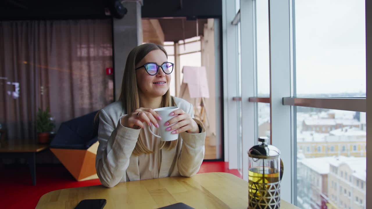 Relaxed lady drinks tea enjoying lunch time rest. Young smiling woman sitting at the table and looking in the window.